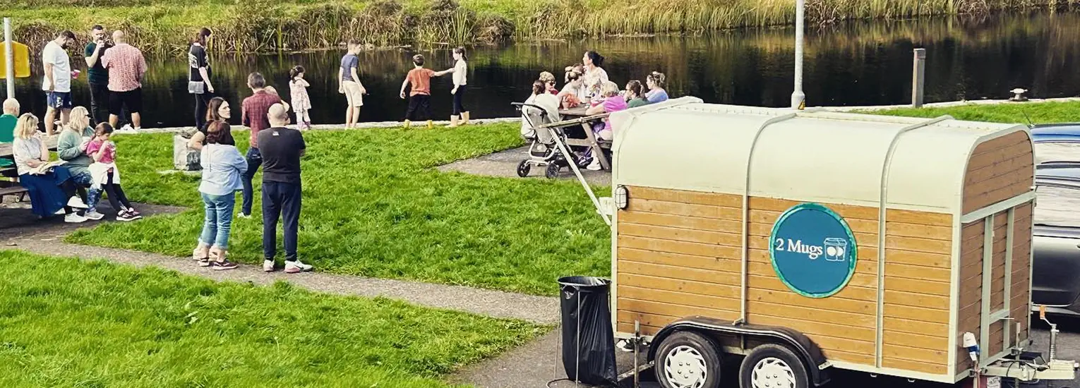 People gather by a riverside on a grassy area, some sitting and some standing, near a small trailer labelled "2 Mugs" that appears to be a mobile coffee stand. The atmosphere is casual and social.