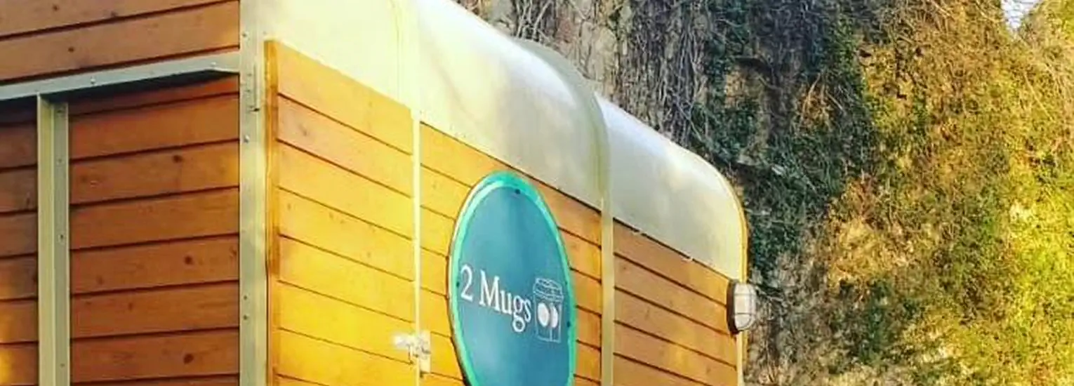 A small wooden coffee stall named "2Mugs" with a blue sign, situated outdoors near a stone wall covered with greenery.