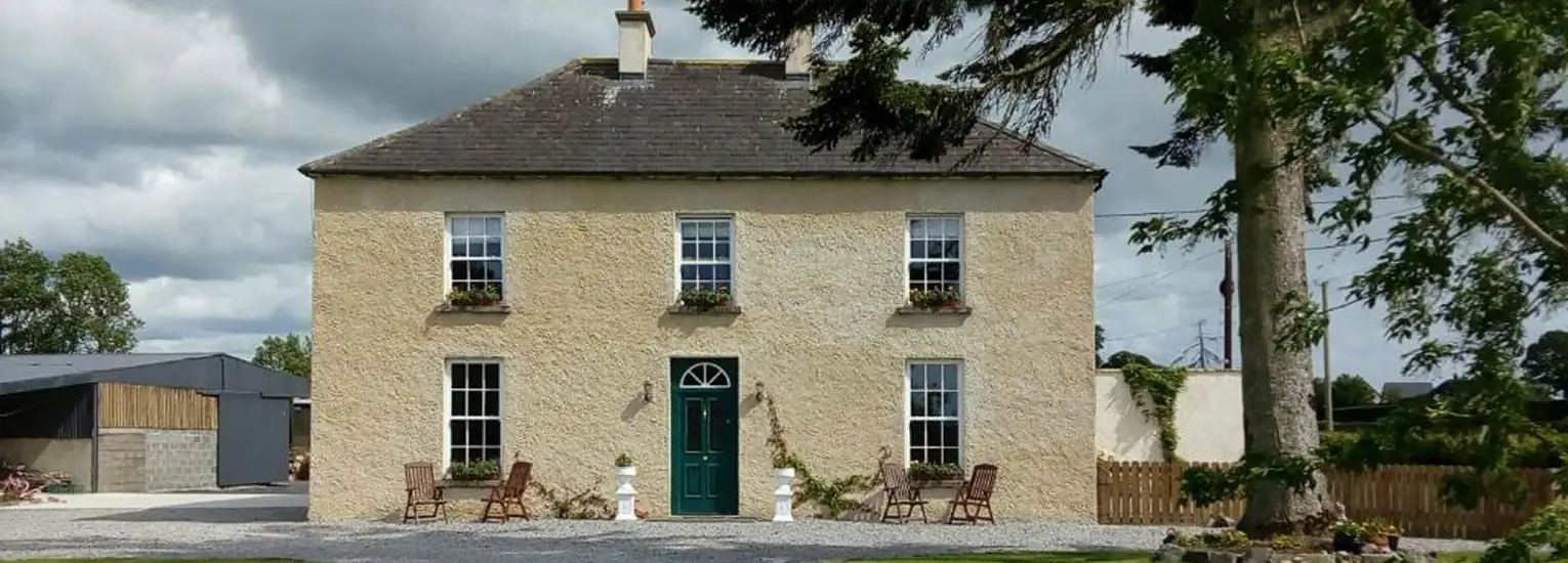 A two-storey country house with a beige exterior, green front door, and four symmetrically placed windows, each with flower boxes. There are chairs near the entrance, trees, and a cloudy sky in the background.