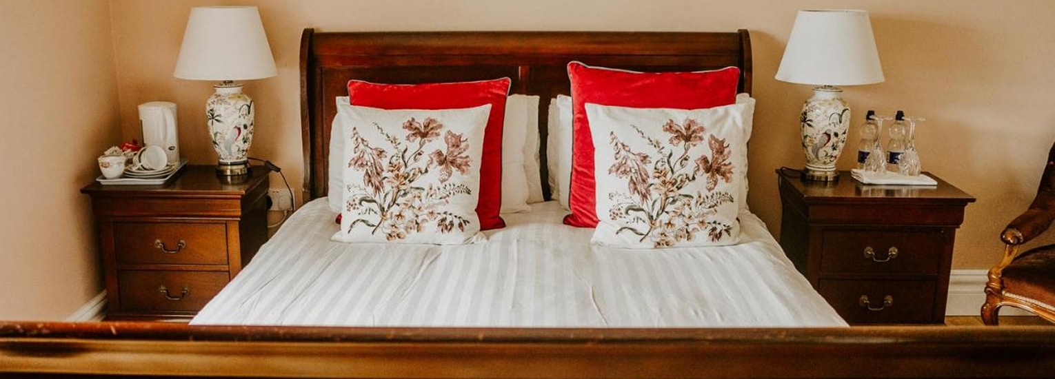 A neatly made bed with white linen, two floral decorative cushions, and two red cushions, flanked by dark wooden bedside tables with matching lamps, water bottles, and a tea set in a cosy, softly lit room.