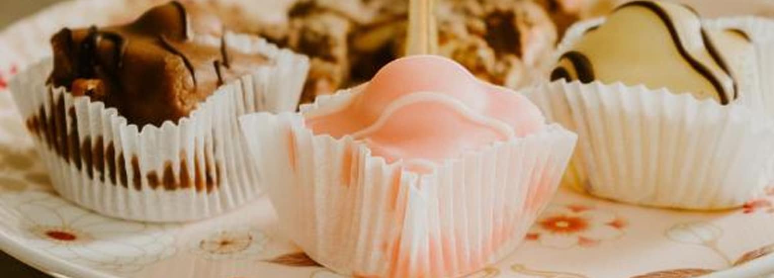 A close-up of assorted chocolates and truffles in paper wrappers on a decorative plate, including pink, white, and brown sweets with different drizzled patterns.