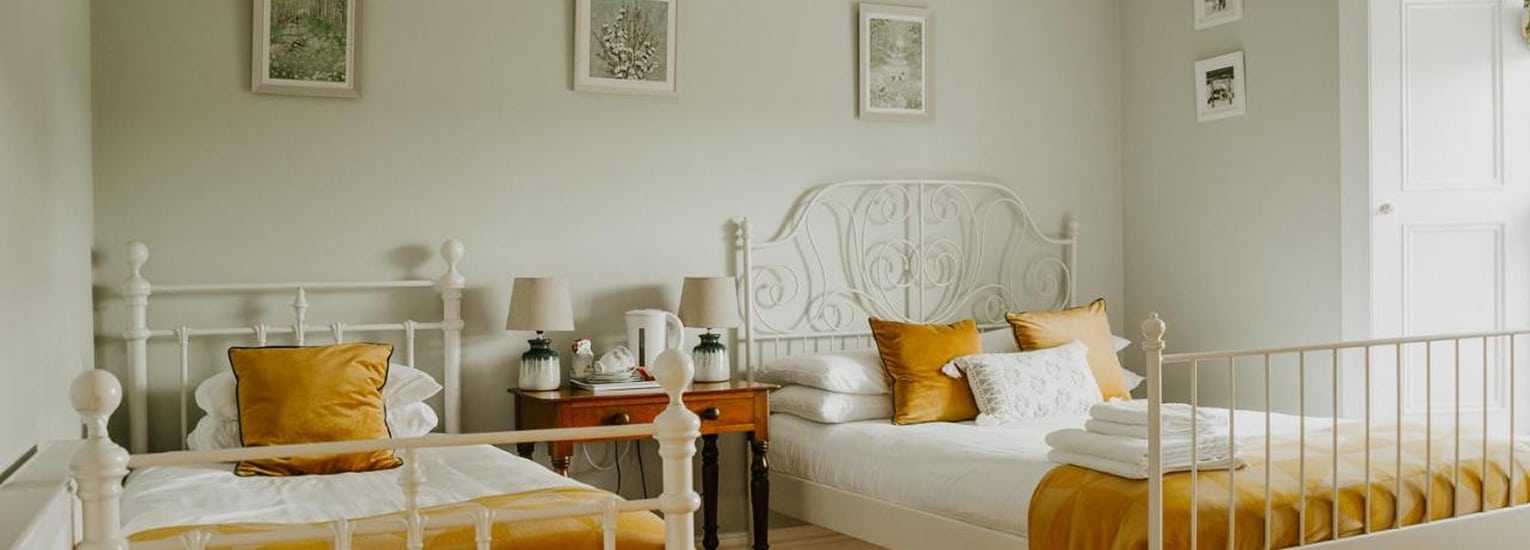 A bright bedroom with two white metal-framed single beds, each with mustard yellow bedding and cushions. A wooden bedside table with lamps sits between the beds. Light-coloured walls feature framed art. Folded towels are on one bed.