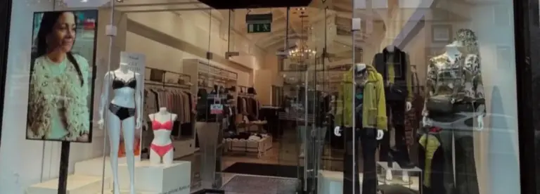 A clothing shop display with two mannequins in lingerie, two mannequins in casual outfits, and a digital screen showing a woman’s portrait. Glass doors reveal more clothing and a chandelier inside.
