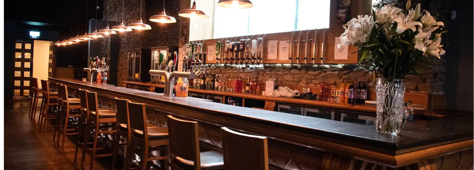 A dimly lit bar with wooden stools lined along a long counter, pendant lights hanging above, a stone wall behind the bar, shelves with bottles, and a vase of white lilies on the counter.