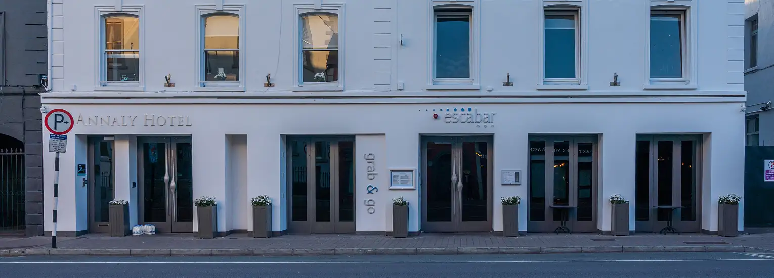 The exterior of a white, modern building with large windows, labelled “Annah Hotel” and “escabar,” featuring glass doors, potted plants, and a no-parking sign on a quiet street.