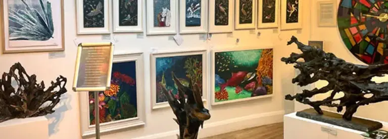 Art gallery with colourful paintings of underwater scenes and coral reefs on the walls, and dark, abstract sculptures displayed on white plinths in the foreground.