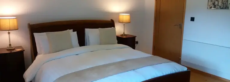 A neatly made bed with white bedding and beige accents sits between two wooden bedside tables with lamps in a bright, minimalist bedroom with wooden flooring and a closed wooden door.