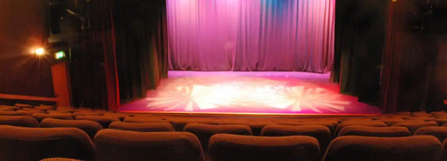 A theatre stage with purple curtains is illuminated by spotlights, viewed from the audience seating area with empty brown chairs in the foreground.