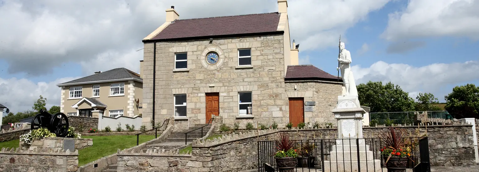 A stone building with a clock above the door stands behind a white statue memorial, surrounded by a stone wall, plants, and a cloudy blue sky. A modern house is visible to the left.