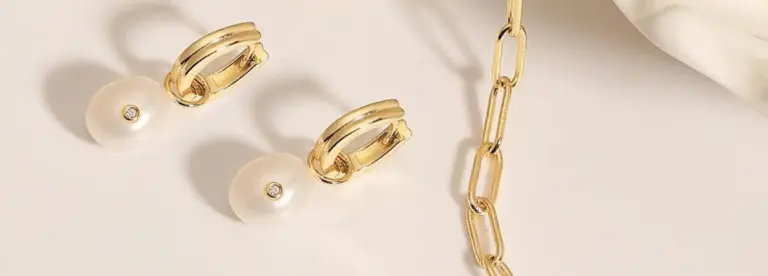 Gold hoop earrings with white pearl and diamond accents are placed beside a gold chain link necklace on a light beige surface. A soft white fabric is visible in the top right corner.