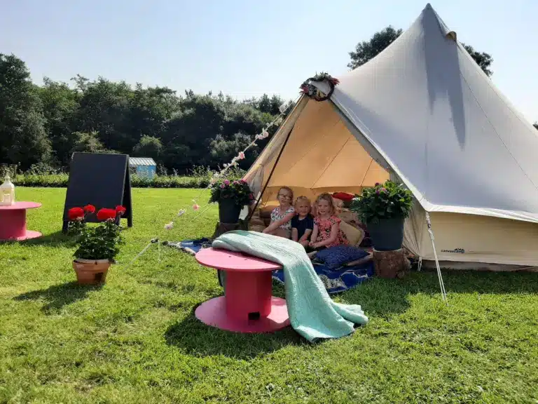 Three children sit inside a cream-coloured bell tent on grass, surrounded by potted flowers and pink tables. A cosy blanket is spread in front, with trees and a chalkboard in the background under a sunny sky.