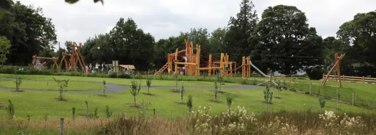 A large wooden playground structure surrounded by grass, trees, and walking paths, with children and adults playing and walking in the park on a cloudy day.