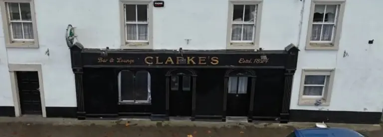 A two-storey white building with a black shopfront sign reading "CLARKE'S Bar & Lounge Estd. 1841", featuring several windows and a parked blue car partially visible at the bottom right.