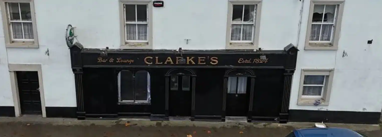 A two-storey white building with a black shopfront sign reading "CLARKE'S Bar & Lounge Estd. 1841", featuring several windows and a parked blue car partially visible at the bottom right.