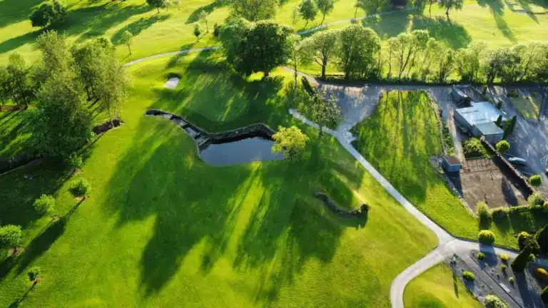 Aerial view of a green golf course with trees casting long shadows, a small pond, winding pathways, and manicured lawns on a sunny day.