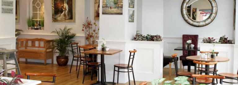 A cosy café interior with wooden floors, round tables, chairs, potted plants, wall art, and a large decorative mirror. The atmosphere is warm, welcoming, and softly lit.
