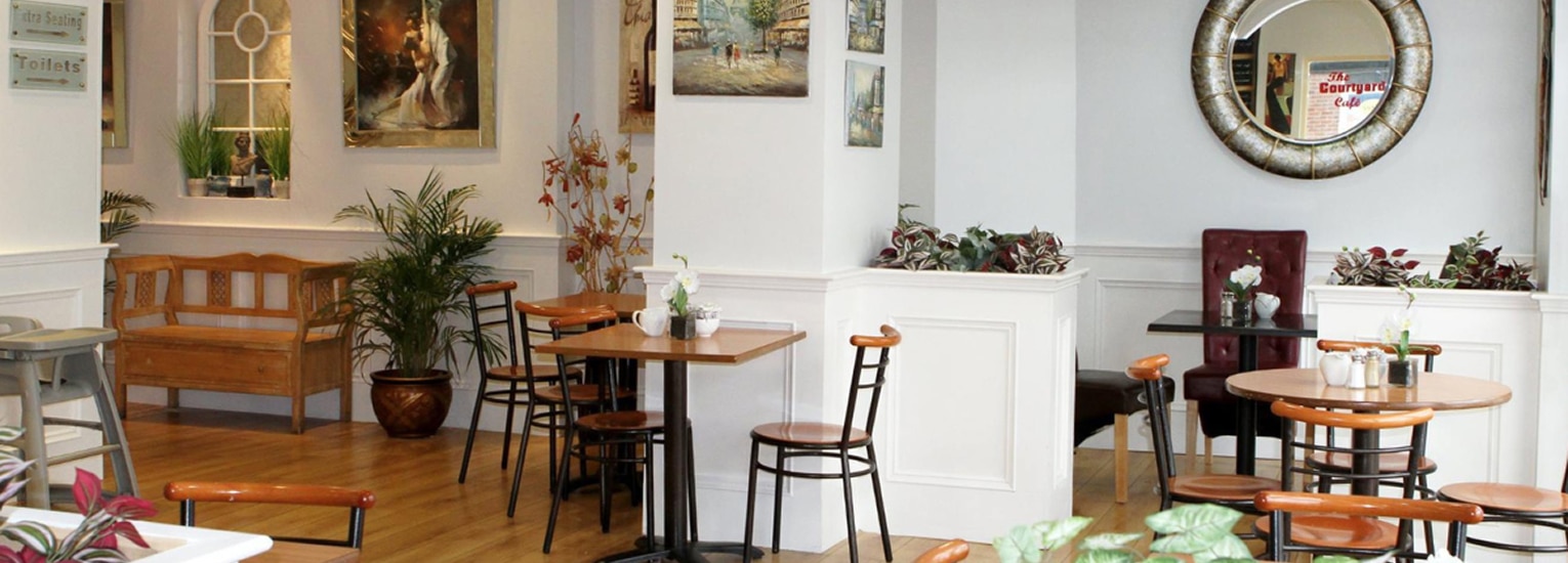 A cosy café interior with wooden floors, round tables, chairs, potted plants, wall art, and a large decorative mirror. The atmosphere is warm, welcoming, and softly lit.