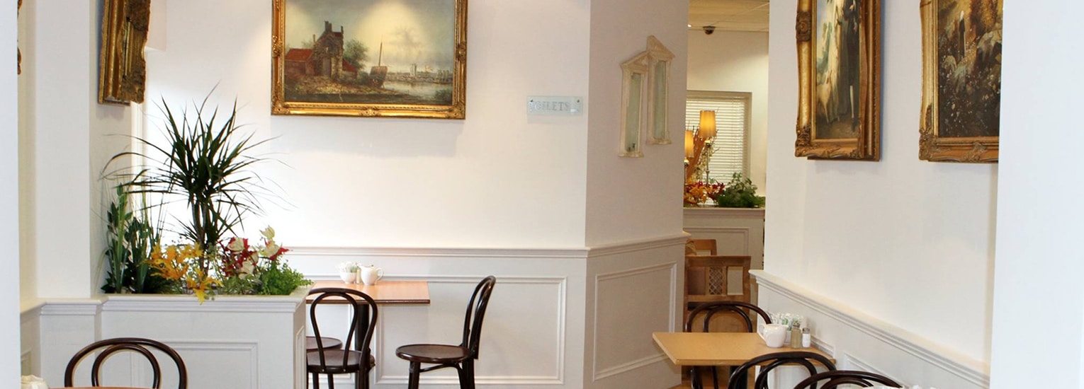 A cosy café with wooden tables and chairs, white walls, framed paintings, and potted plants creating a welcoming atmosphere.
