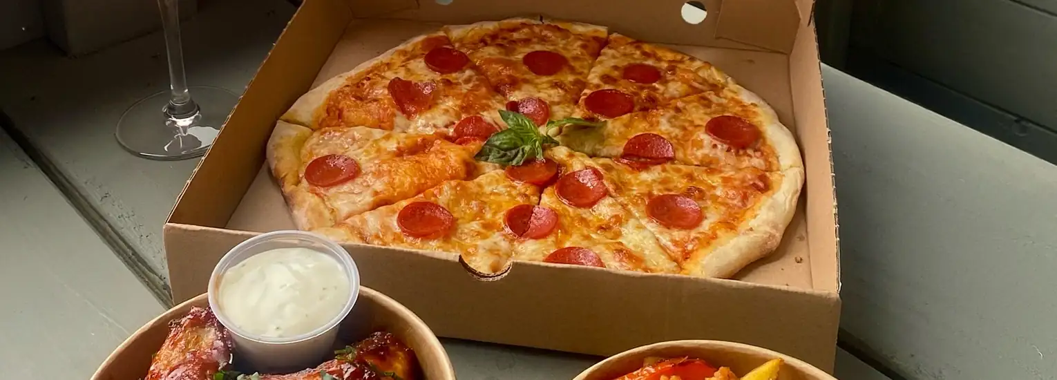 A pepperoni pizza in an open cardboard box is topped with a sprig of basil. In front of it, there are two bowls: one with chicken wings and dip, the other with chips and sauce.