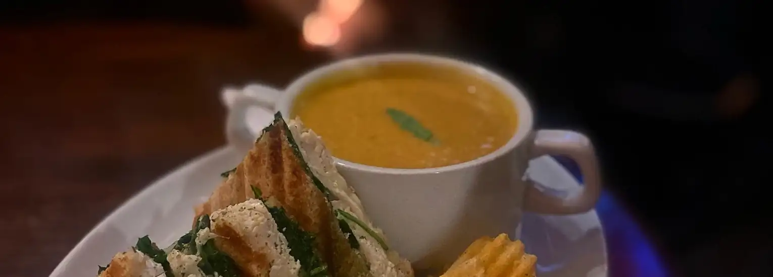 A bowl of creamy soup garnished with a fresh basil leaf, served with a toasted sandwich and crinkle-cut crisps on a white plate.