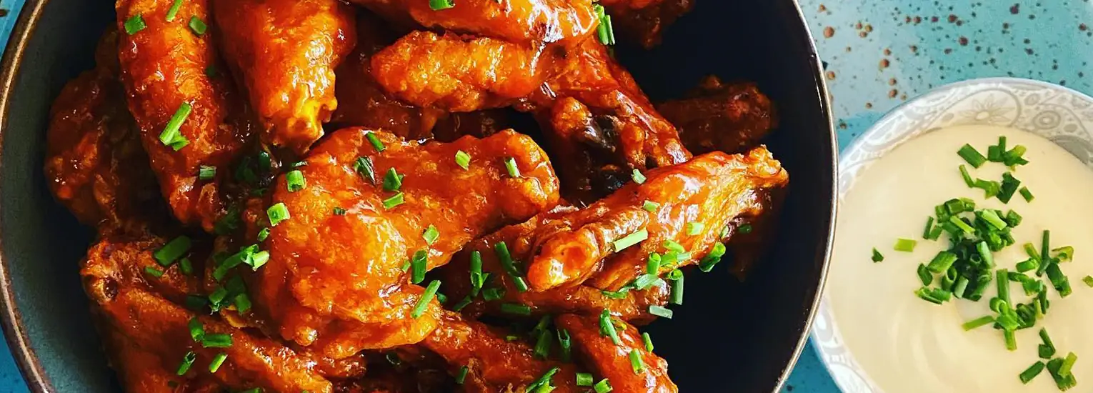 A bowl of saucy buffalo chicken wings garnished with chopped chives, next to a small dish of creamy dipping sauce topped with more chives, on a blue speckled surface.