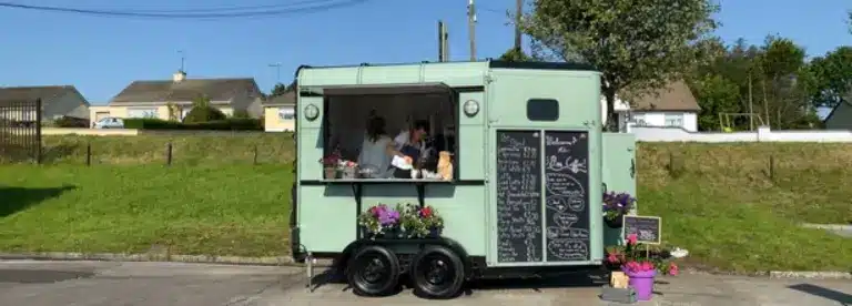 A mint green food truck with a large serving window and chalkboard menu is parked by a grassy area. Two people are inside serving customers. Potted flowers decorate the front of the lorry. Houses and trees are in the background.