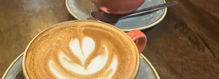 Two ceramic cups on saucers sit on a wooden table. The front cup contains a latte with a leaf-shaped foam design, and the other cup in the background has a spoon resting on its saucer.