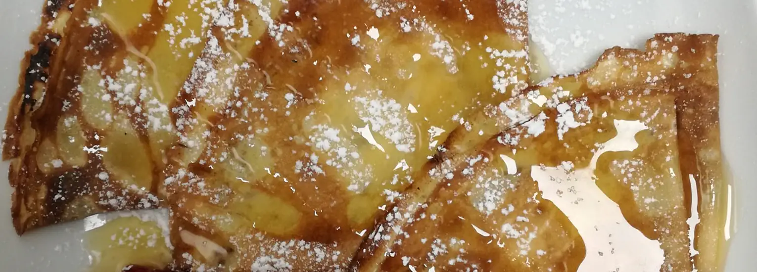 Several golden-brown crêpes are drizzled with syrup and sprinkled with icing sugar, served on a white plate.