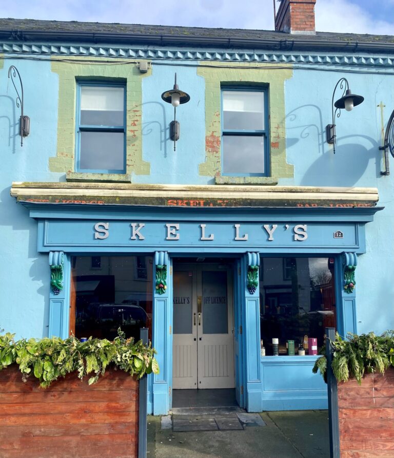 A blue-painted building with green trim, featuring a sign that reads "SKELLY'S" above double doors. There are outdoor lights, windows, and planters with greenery in front.