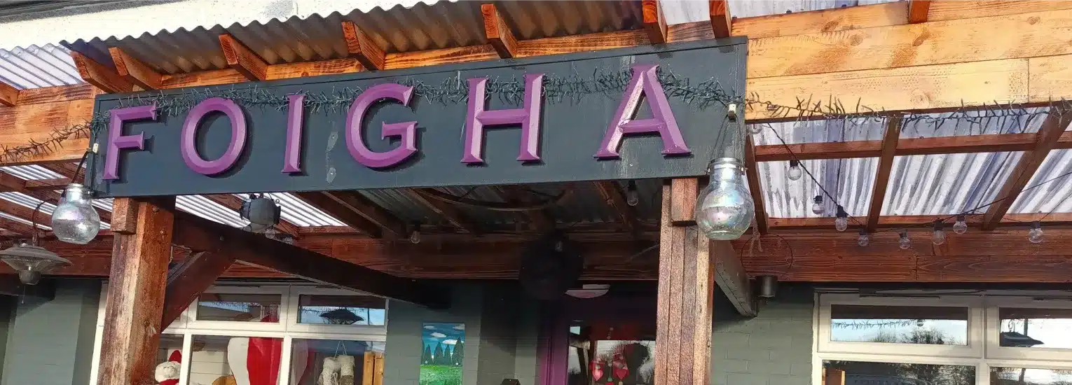 A wooden sign with large purple letters spelling "FOIGHA" hangs above the entrance of a building with a clear corrugated roof and fairy lights.
