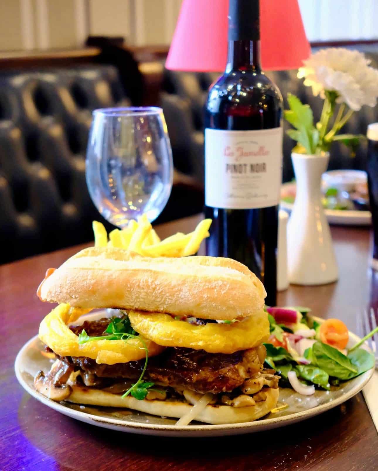 A gourmet sandwich with fried onion rings, meat, and greens is served with chips and salad on a plate. A bottle of red wine and a wine glass are on the table, with a flower vase and lamp in the background.