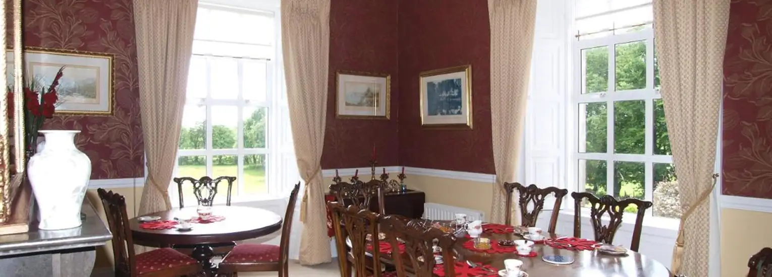 Bright dining room with large windows, wooden tables and chairs, cream curtains, red walls with framed art, and a tea set arranged on the main table. Greenery is visible outside.