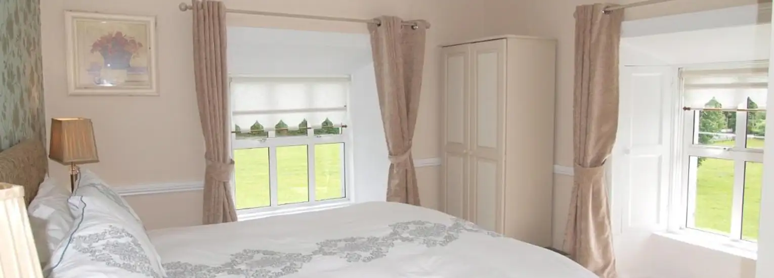 Bright bedroom with a double bed, white bedlinen, two large windows with beige curtains, a wardrobe, and a framed flower picture on the wall; green landscape visible outside.