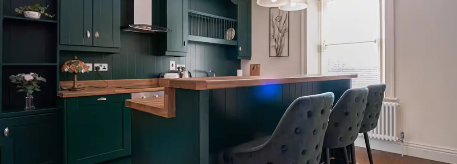A modern kitchen with dark green cupboards, wooden worktops, a breakfast bar with three upholstered chairs, pendant lights above, and a large window letting in natural light.