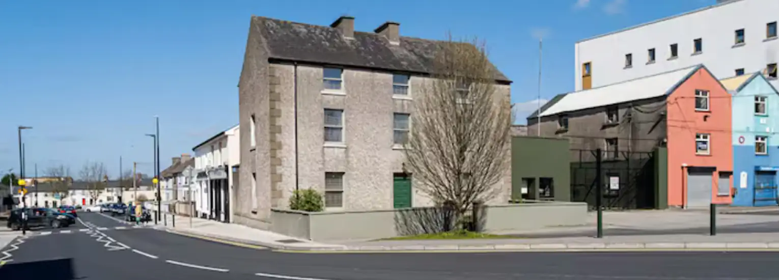 A quiet urban street features a large grey brick building, colourful houses, and a leafless tree in front. The sky is clear and blue, and there are no people visible.