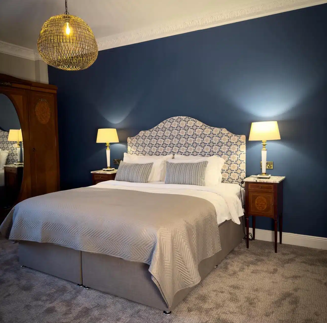 A stylish bedroom with a large bed, patterned headboard, beige bedding, two bedside tables with lamps, a wooden wardrobe with a mirror, blue feature wall, and a woven pendant light hanging from the ceiling.
