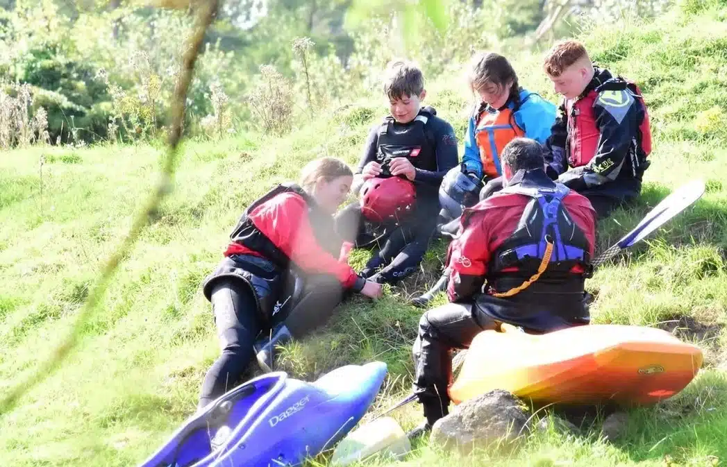 Five children wearing wetsuits and life jackets sit on a grassy slope with two kayaks beside them, appearing to take a break and talk together outdoors on a sunny day.