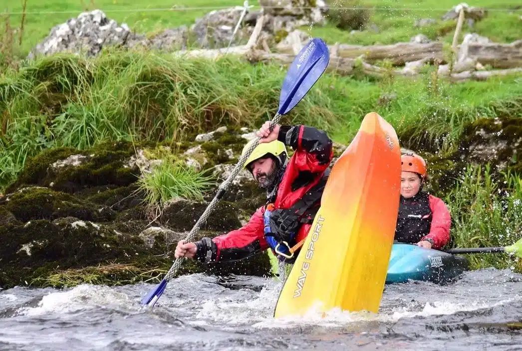 Two people kayaking in a river; the person in front is tipping their orange and yellow kayak sideways whilst paddling, and the person behind is smiling, both wearing helmets and life jackets. Lush green grass and rocks in the background.