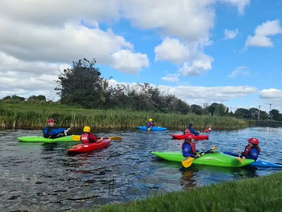 A group of six people in colourful kayaks paddle on a calm river, surrounded by green grass and bushes under a blue sky with scattered clouds. All are wearing helmets and life jackets.