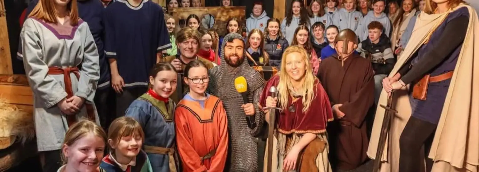A large group of people, many dressed in medieval-style costumes, pose indoors for a photo. A woman holding a microphone stands in front, with others smiling and wearing robes, tunics, or chain mail.