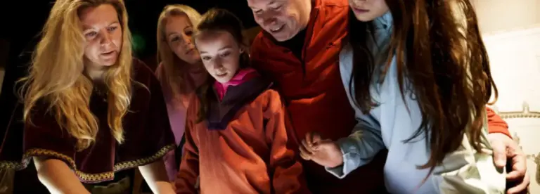 A group of five people, including adults and children, gather closely around a table, looking intently at something on its surface, illuminated by a bright light. They appear engaged and focussed.