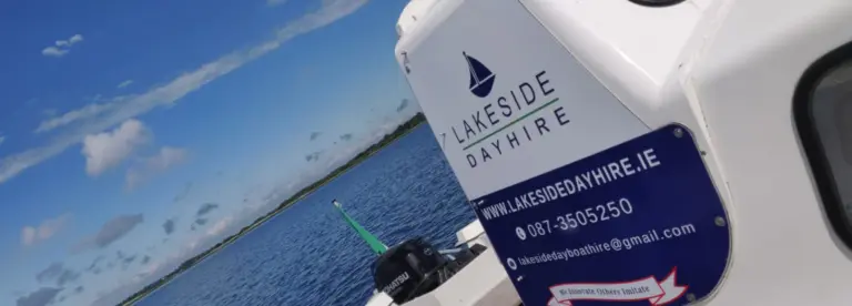 A close-up of a boat with a "Lakeside Day Hire" sign on the side, floating on a lake under a blue sky with scattered clouds. Water and distant shoreline are visible in the background.