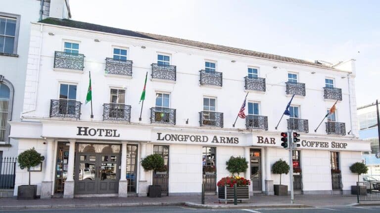 A white three-storey building with decorative balconies, flags, and signs reading "Hotel Longford Arms," "Bar," and "Coffee Shop." There are potted trees and flowers outside the entrance.