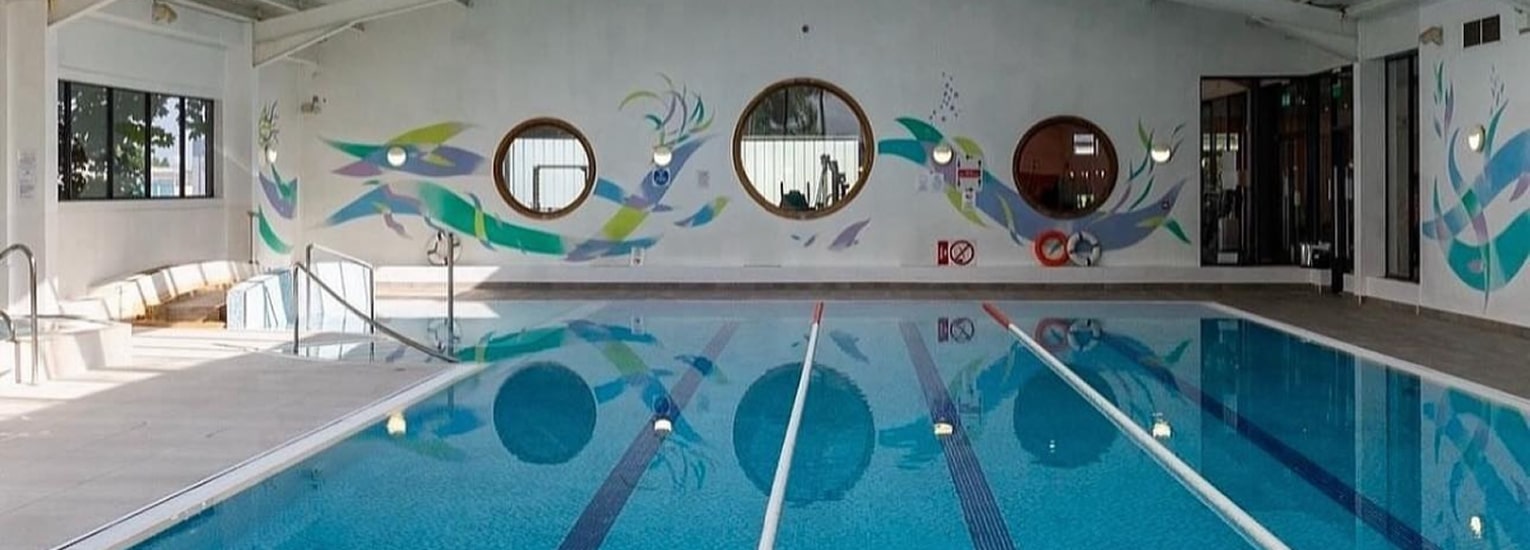 Indoor swimming pool with two swimming lanes, circular windows on the wall, and colourful fish murals. There are loungers on the left and natural light streaming in from large windows.