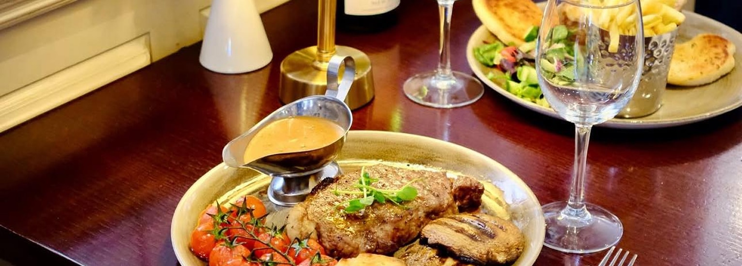 A plated steak meal with grilled tomatoes, mushrooms, and sauce is set on a wooden table beside a glass of wine, empty wine glasses, a candle, and another plate with chips, salad, and bread in the background.