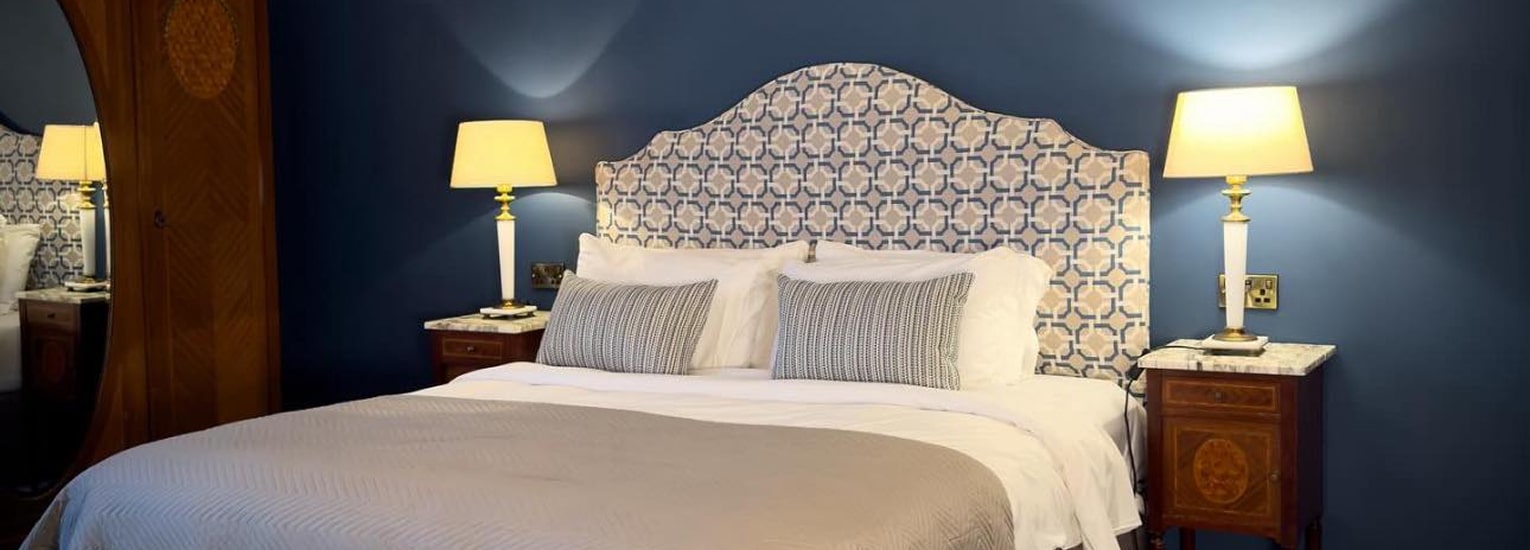A cosy bedroom with a large bed, patterned headboard, two pillows, and a beige blanket. The bed is flanked by wooden bedside tables, each with a lit lamp, against a dark blue wall.