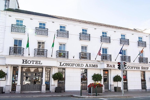 White three-storey building labelled “Longford Arms Hotel” with balconies, flags, large windows, and shopfronts for a bar and coffee shop; potted plants and a traffic light are visible at the front.