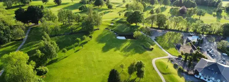 Aerial view of a lush green golf course with sand bunkers, trees, winding paths, and a clubhouse with parked cars on a sunny day.
