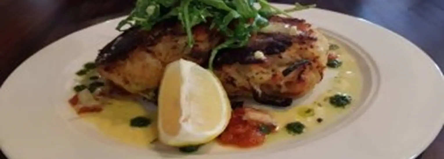 A grilled chicken breast garnished with fresh greens and placed on a creamy sauce with herbs, served with a lemon wedge on a white plate.