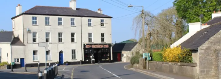 A quiet village street curves past a large, pale three-storey building with a shop on the ground floor, surrounded by stone buildings, greenery, and a clear blue sky.